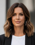 A woman with shoulder-length wavy brown hair wearing a black blazer over a white top, accessorized with large silver hoop earrings, shown in a close-up portrait.