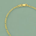 A close-up of a gold Figaro chain bracelet with a lobster clasp, displayed on a textured sage green background.