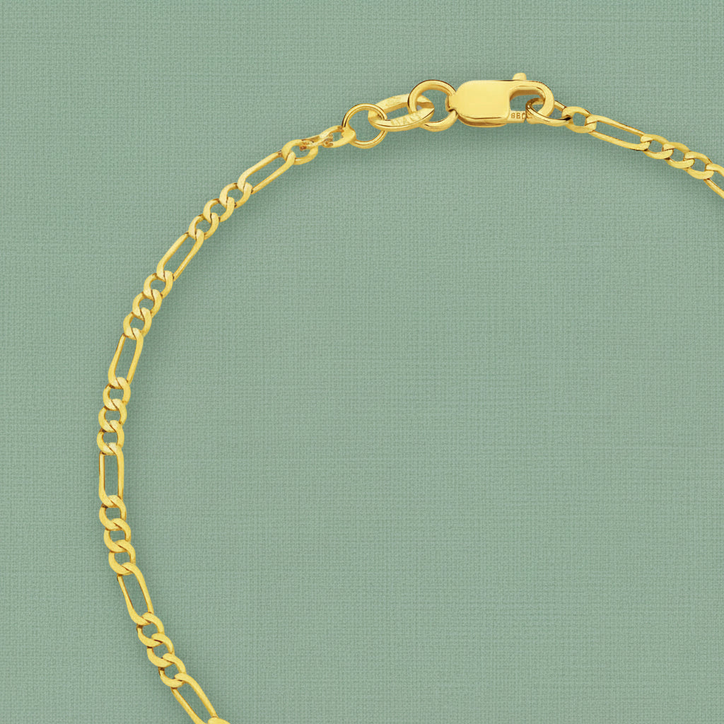 A close-up of a gold Figaro chain bracelet with a lobster clasp, displayed on a textured sage green background.