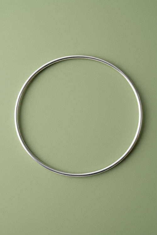 A thin 2mm, polished silver metal circular  bangle laid flat on a solid sage green background.