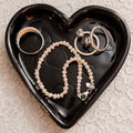 Heart-shaped black jewelry dish with rings and a bracelet on a lace background