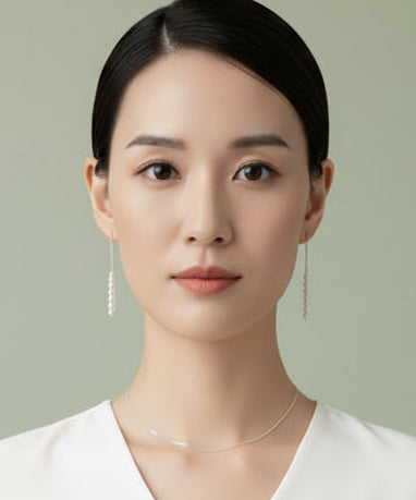 A close-up of a woman wearing long, dangling silver thread earrings with a small cluster of freshwater pearls at the bottom, paired with a delicate silver chain necklace.