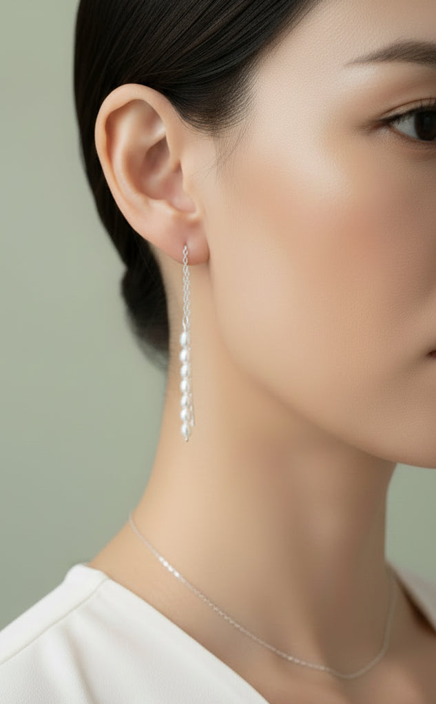A close-up of a person's ear wearing a long silver thread earring with seven small freshwater pearls hanging vertically