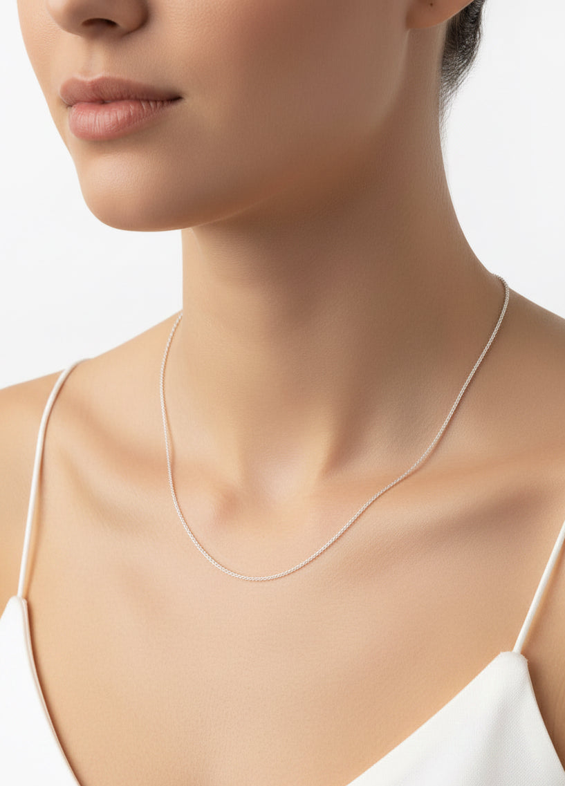 A close-up of a model's neck and upper chest wearing a delicate, thin silver cable chain necklace with a white camisole top against a clean white background.
