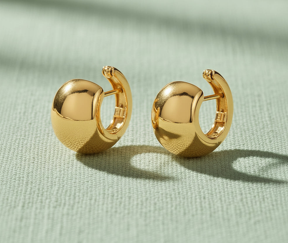 A pair of polished gold plated silver chunky huggie hoop earrings with a textured lower half, displayed on a textured beige fabric background.